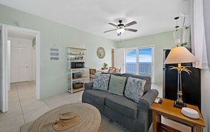 A living room with a grey sofa, a ceiling fan, a lamp, and a view of the outside.