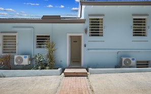A building with a light blue exterior, a front door, two air conditioning units, and a small garden area with plants.