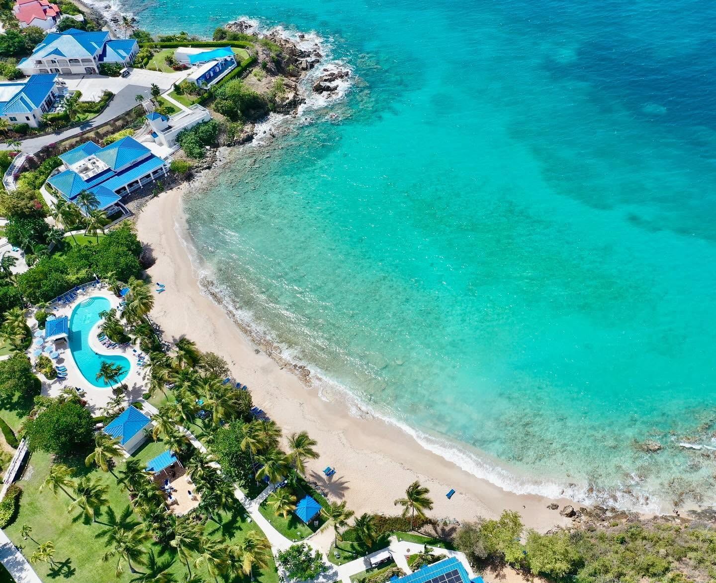 An aerial view of a coastal resort with a clear turquoise sea, a sandy beach, and a swimming pool.