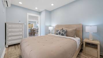 A bedroom with a large bed, bedside lamps, a dresser, and a view into the dining area.