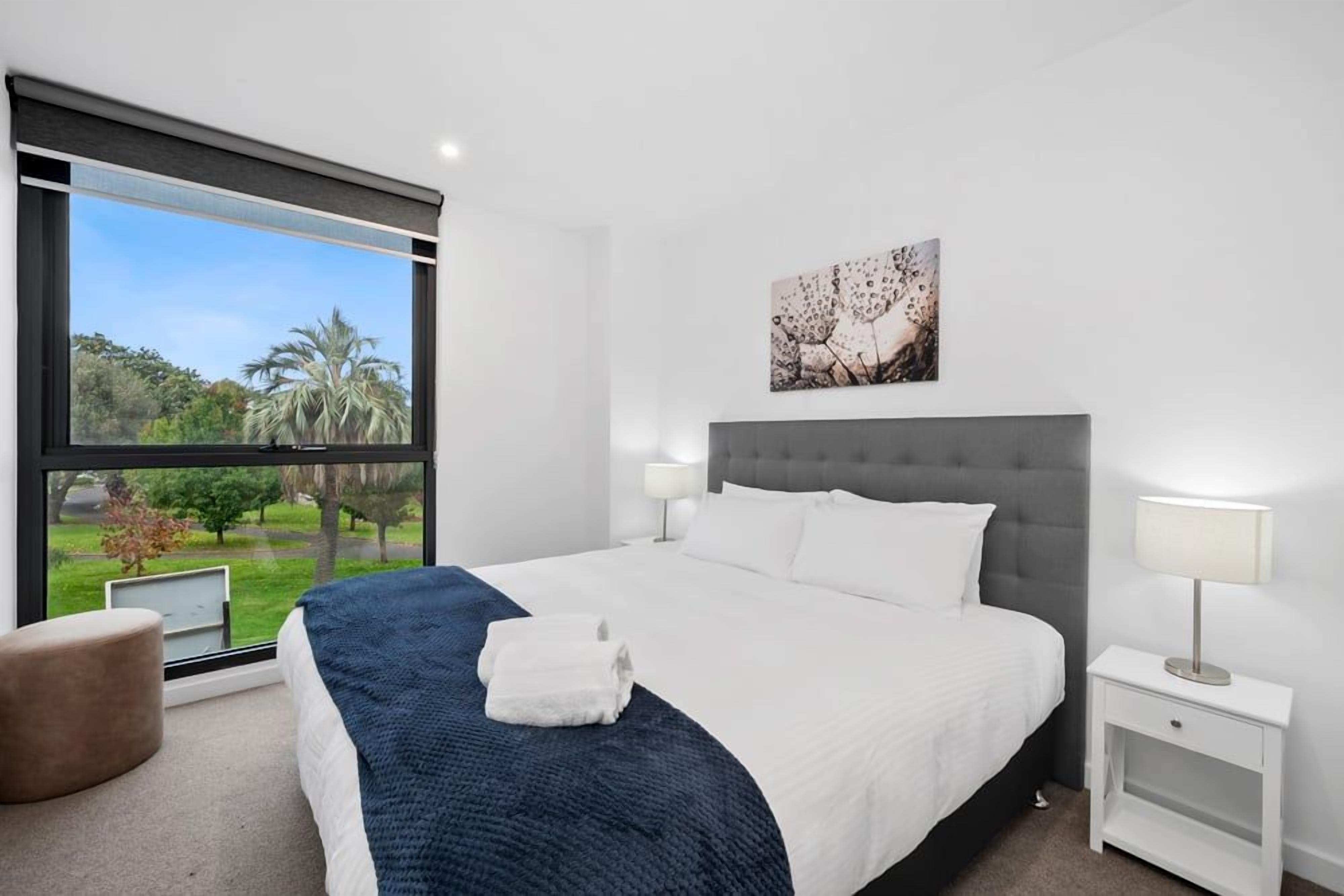 A modern hotel room with a large bed, a view of greenery outside, a bedside table with a lamp, and a painting on the wall.