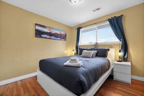 A bedroom with a bed, a nightstand, a window with curtains, a painting on the wall, and a ceiling light.