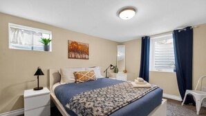 Una camera da letto con un letto, un comodino, una lampada, una finestra con tende, una pianta e un quadro appeso al muro.