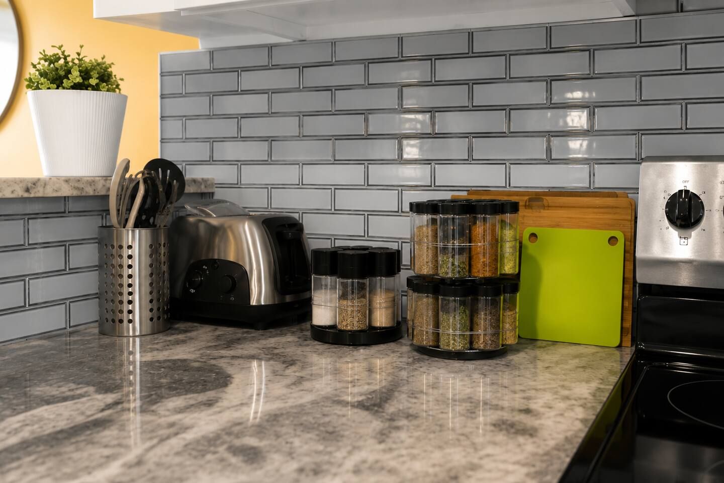 Una cocina con encimera de granito, un tostador, una tabla de cortar verde, frascos de especias y una maceta con plantas verdes.