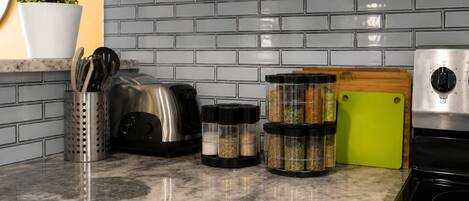 Una cocina con encimera de granito, un tostador, una tabla de cortar verde, frascos de especias y una maceta con plantas verdes.