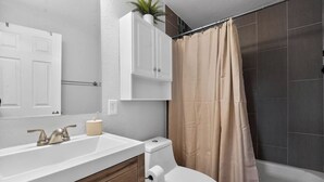 Un bagno con un water bianco, un lavandino con dispenser di sapone color beige, uno specchio e una tenda per la doccia.