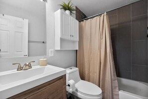 A bathroom with a white toilet, a sink with a beige soap dispenser, a mirror, and a shower curtain. - Hot Tub + Fire Pit + Movie Projector+ BBQ - Near DT Denver (Thornton)
