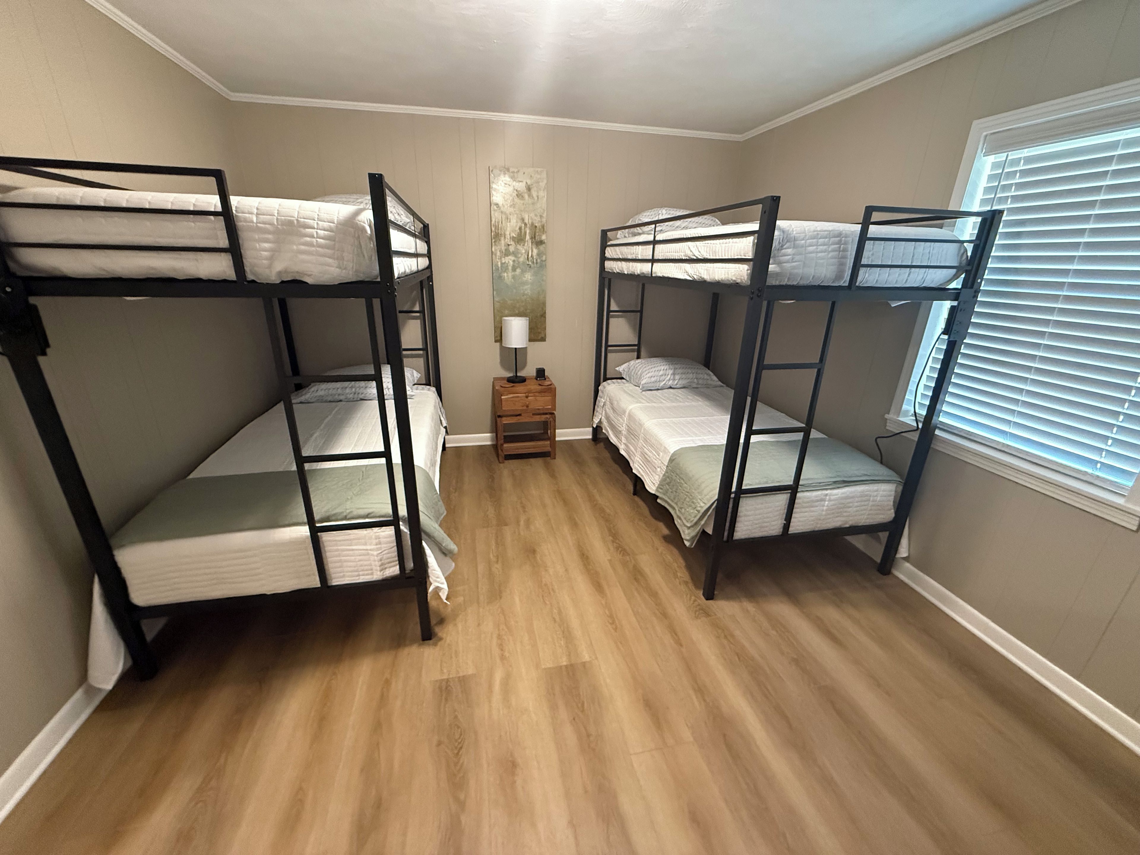 Une chambre avec des lits superposés, une petite table de chevet en bois, une lampe et une fenêtre avec des stores.