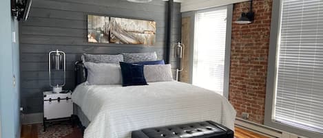 Une chambre à coucher moderne avec un grand lit, une commode, un banc et un tableau représentant un paysage urbain accroché au mur.