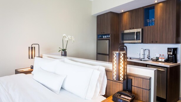 Quarto de hotel com uma cama, mesa de cabeceira com luminária, pequena cozinha equipada com micro-ondas, geladeira e máquina de café.