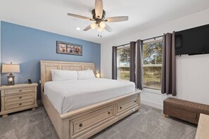 Een slaapkamer met een bed, nachtkastjes, een plafondventilator, een raam met gordijnen en een aan de muur bevestigde televisie.