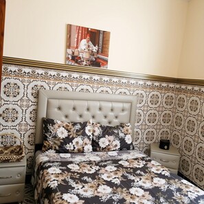 Een slaapkamer met een gestapeld hoofdbord, bloemenbeddengoed, behang met een patroon, een nachtkastje en een ingelijst foto op de muur.