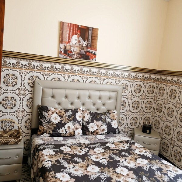 Een slaapkamer met een gestapeld hoofdbord, bloemenbeddengoed, behang met een patroon, een nachtkastje en een ingelijst foto op de muur.