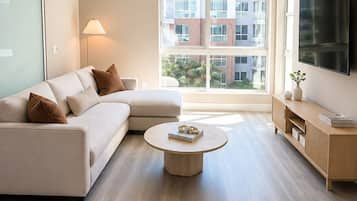 Un salon moderne avec un canapé d’angle, une table basse ronde, un téléviseur et une grande fenêtre donnant sur des immeubles.