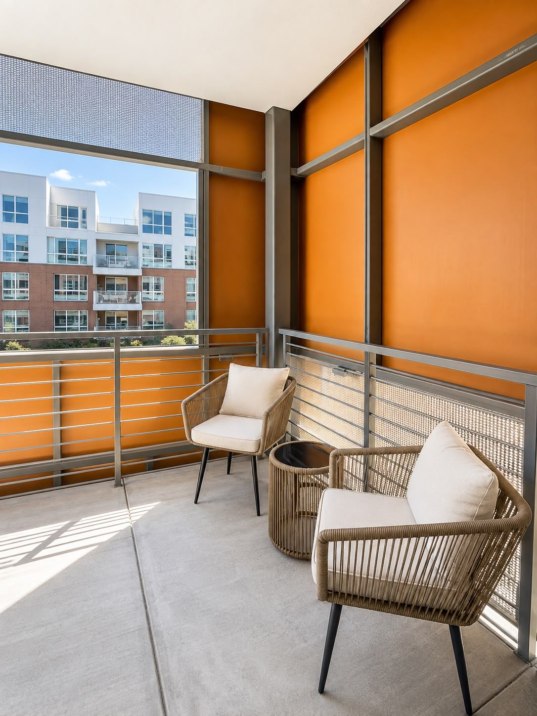 Un balcon avec deux fauteuils en osier et une petite table ronde, donnant sur un immeuble doté de nombreuses fenêtres.