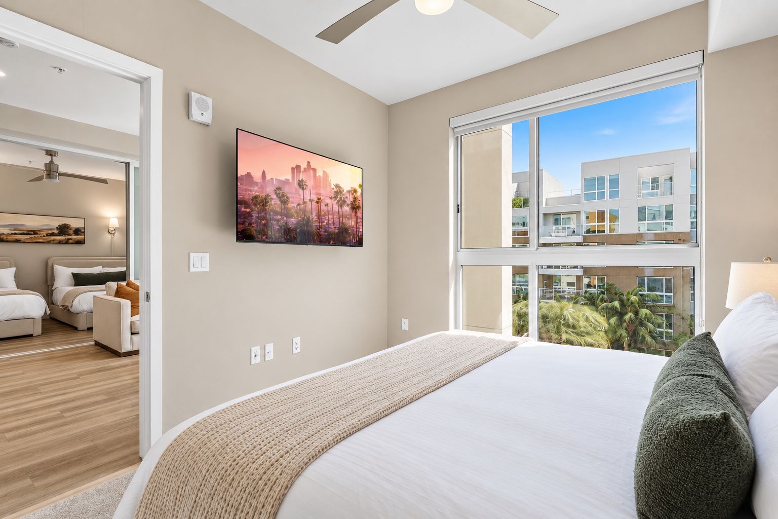 A modern hotel room with a large bed, a cityscape painting, and a view of a building through the window.