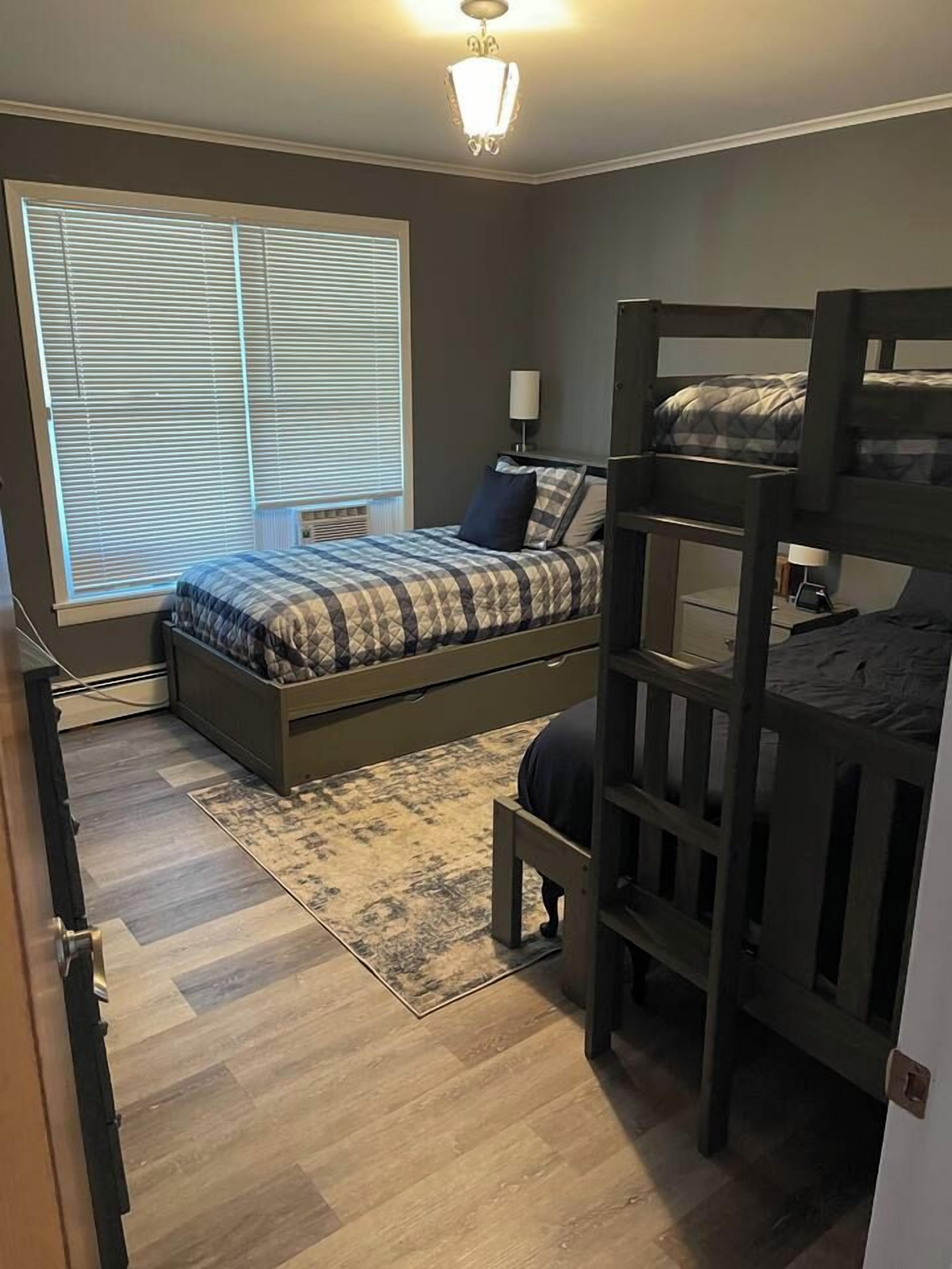 A bedroom with a bunk bed, a single bed, a window with blinds, a bedside lamp, and a rug.