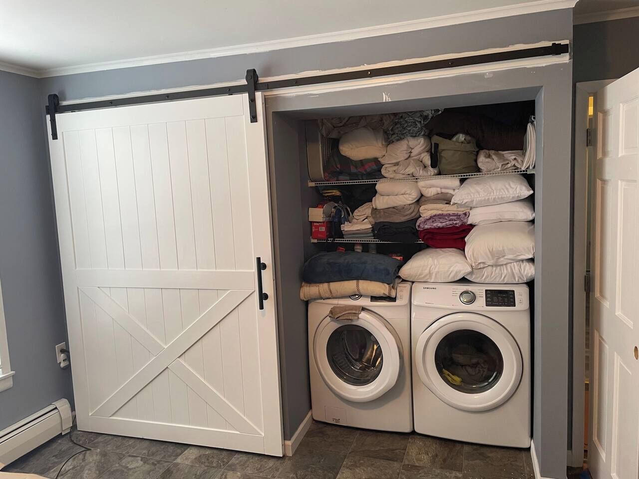 Une buanderie avec une porte blanche, deux machines à laver et une étagère où sont pliés des vêtements et du linge de maison.