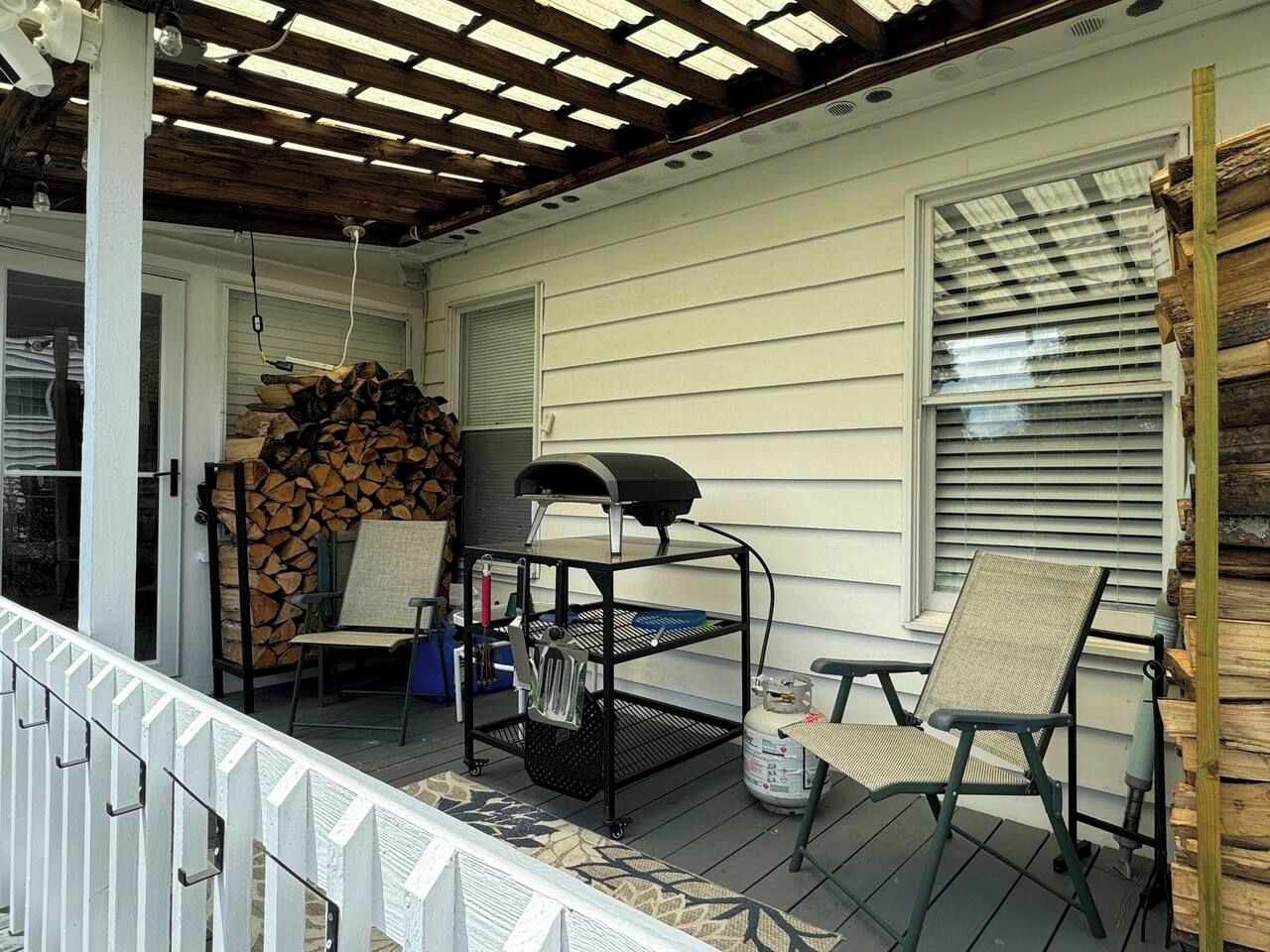 A covered porch with a grill, chairs, and a stack of firewood.