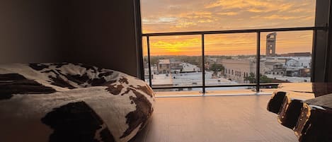 A room with a large window overlooking a cityscape at sunset, a wooden floor, and a cowhide-patterned pillow.