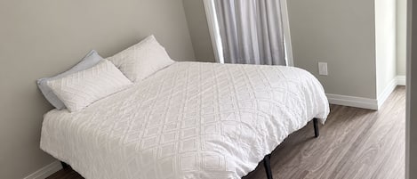 A bed with a white quilt and pillows, a wooden floor, and a window with curtains.