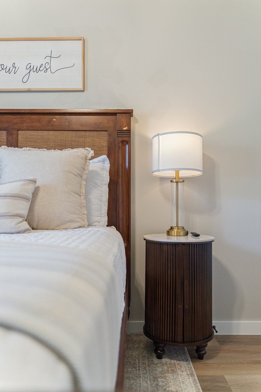 A neatly made bed with white linens and a headboard, a bedside lamp on a wooden nightstand, and a framed sign on the wall.