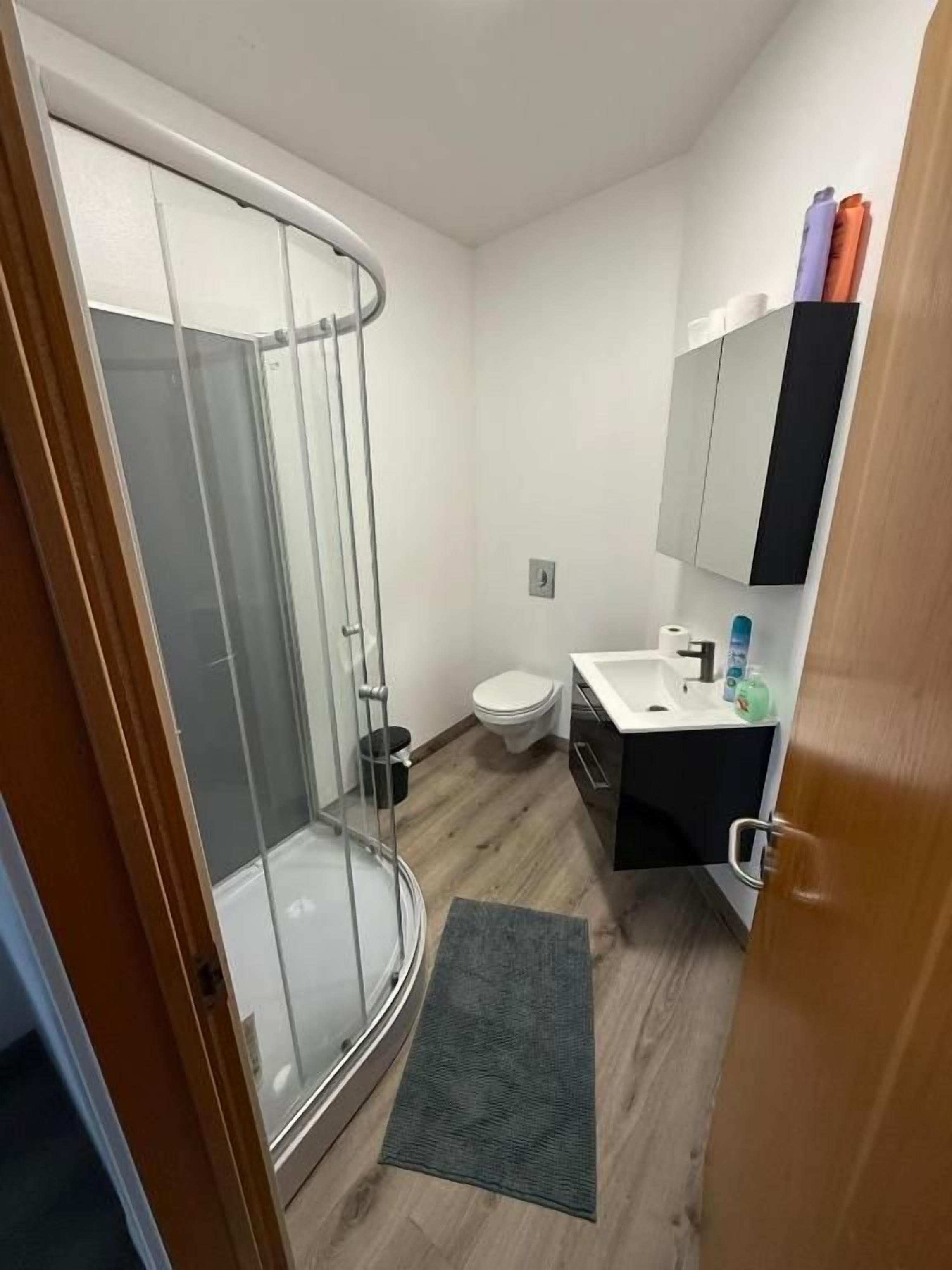 Ein kleines Badezimmer mit Glasdusche, Toilette, Waschbecken mit Spiegel und einem kleinen Schrank.