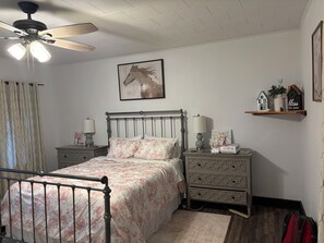 Ein Schlafzimmer mit einem Bett, Nachttischen, einer Kommode, einem Deckenventilator, einem Wandbild und einem Regal mit Dekoration.