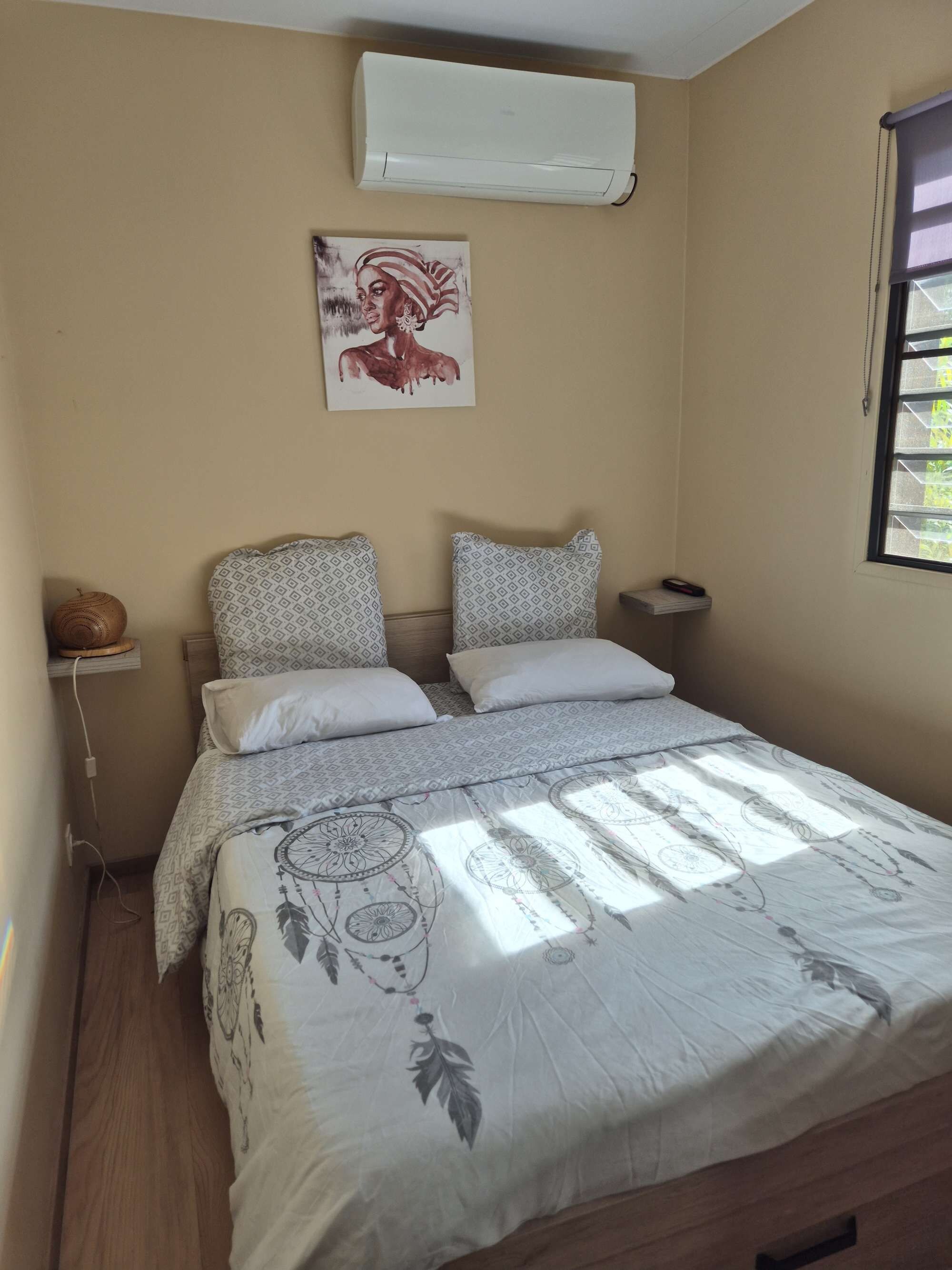 A bedroom with a bed, patterned pillows, a wall-mounted air conditioner, a painting, and a window with blinds.