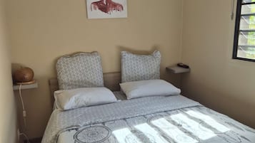 A bedroom with a bed, patterned pillows, a wall-mounted air conditioner, a painting, and a window with blinds.