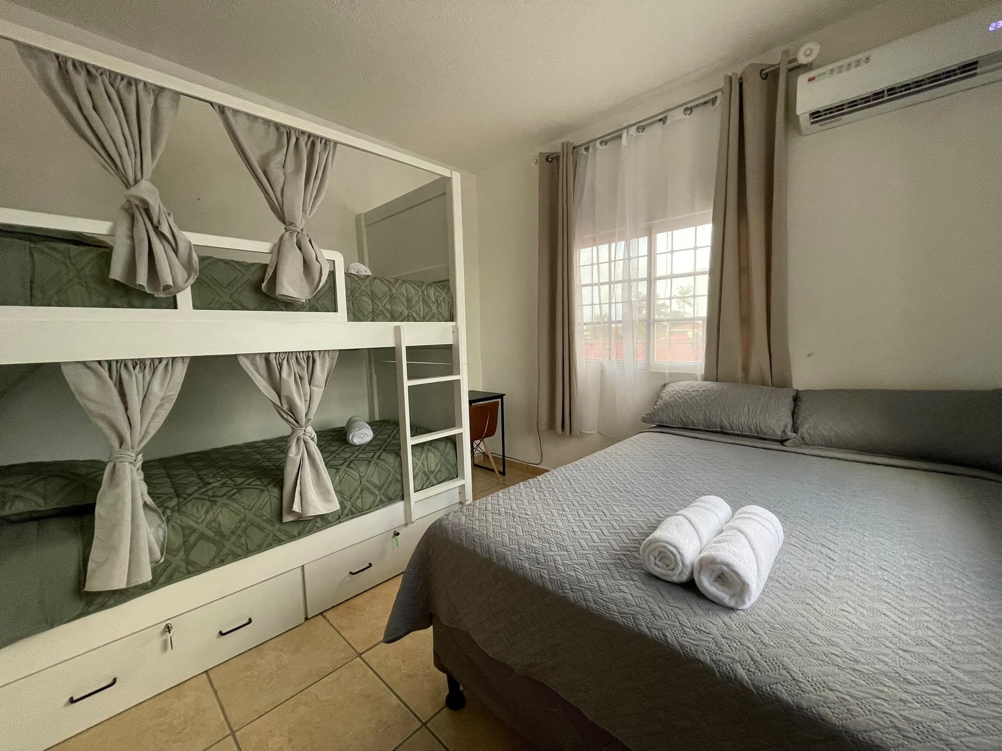 Habitación de hotel con literas, una cama individual, una silla y un ventanal con cortinas.