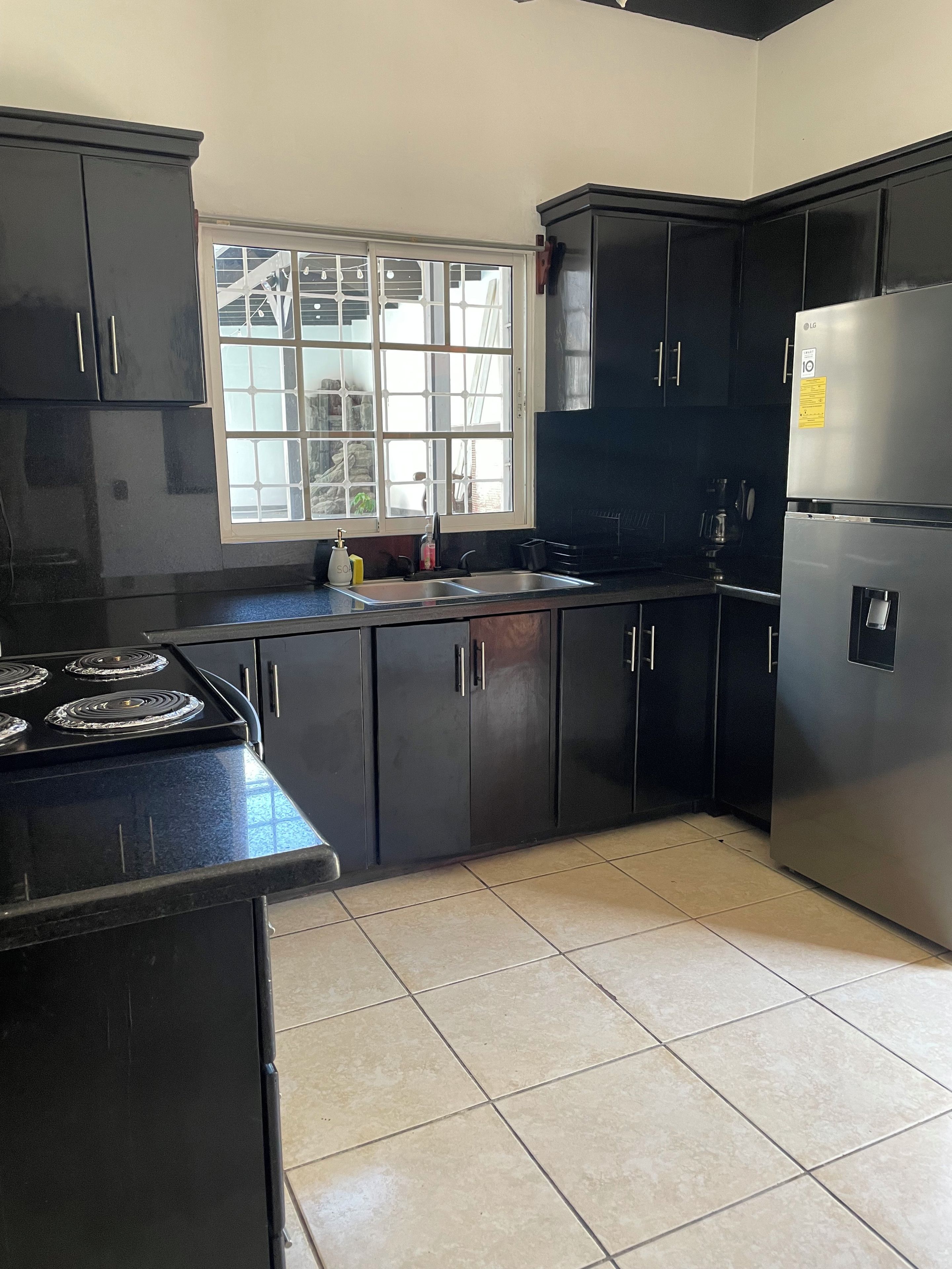 A kitchen with black cabinets, a stainless steel refrigerator, a double sink, and a window with a view.