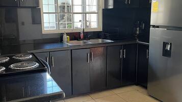 A kitchen with black cabinets, a stainless steel refrigerator, a double sink, and a window with a view.