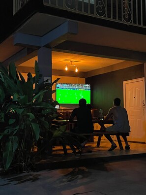 A room with a television displaying a sports game, a dining table, and a potted plant.