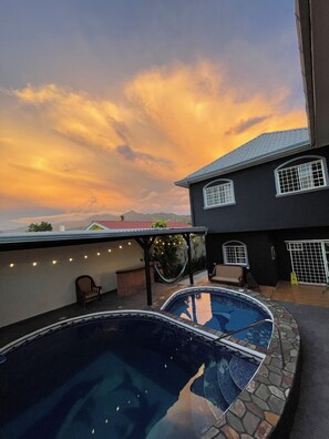 Un área de piscina en la azotea con dos piscinas, una pérgola con luces colgantes y vistas al atardecer.