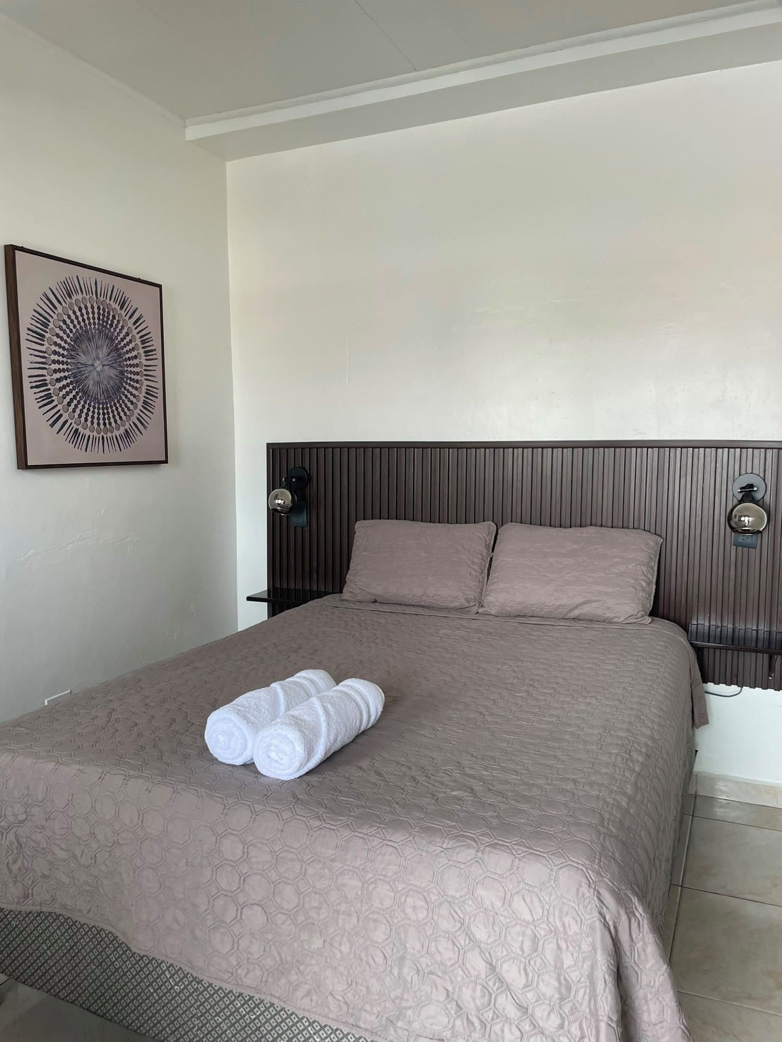 A hotel room with a large bed, two pillows, a headboard with vertical lines, a wall-mounted light fixture, and a framed abstract artwork.