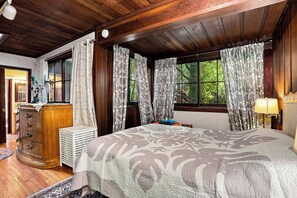 A bedroom with a large bed, wooden ceiling, patterned curtains, a dresser, and a radiator.