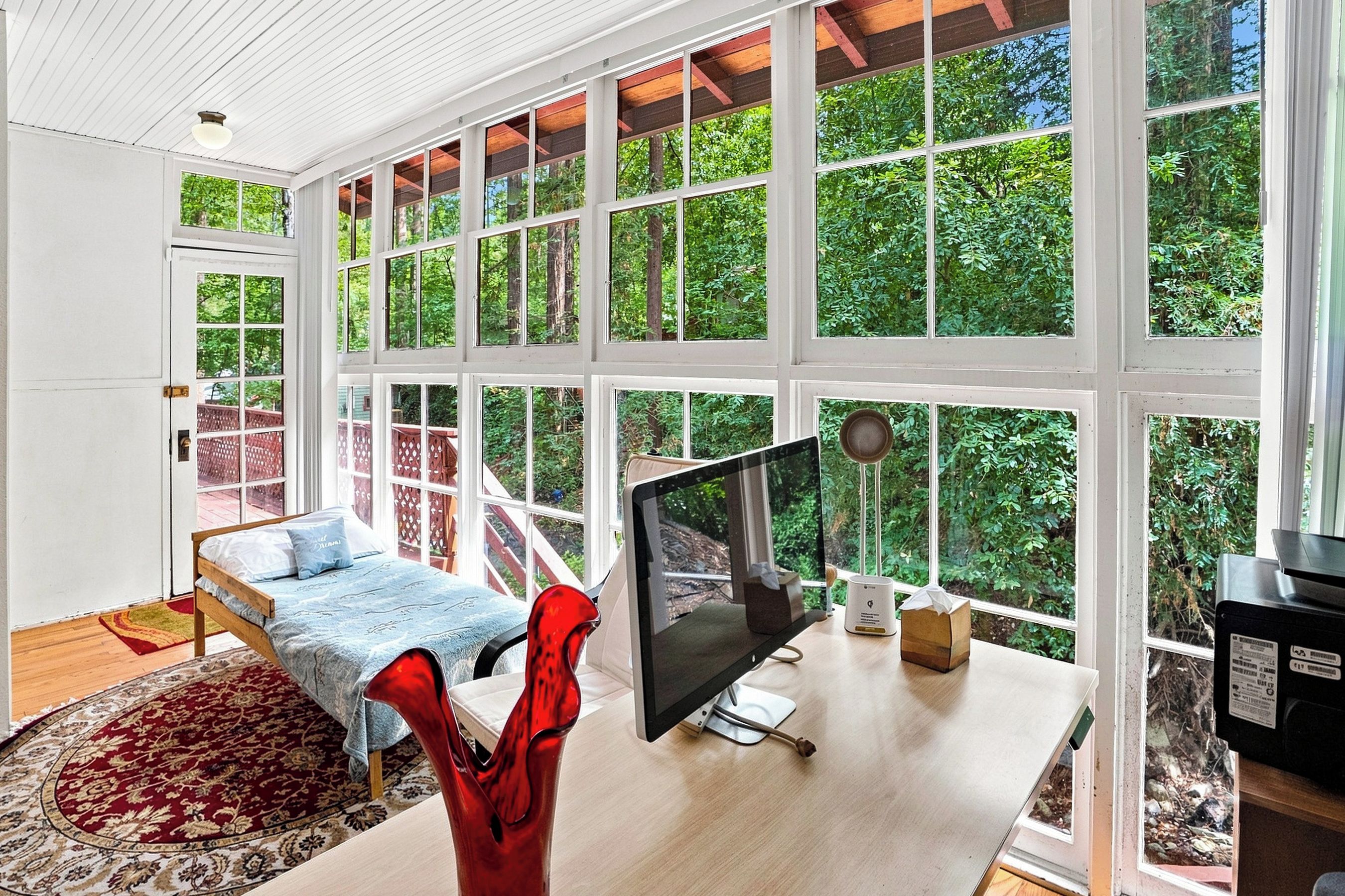 A room with a bed, a desk with a computer, and large windows.