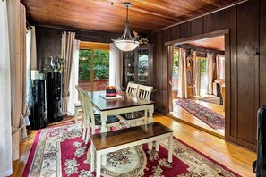 Un comedor con mesa de madera, sillas y una lámpara de araña. Hay una ventana con cortinas y vistas a zonas verdes.