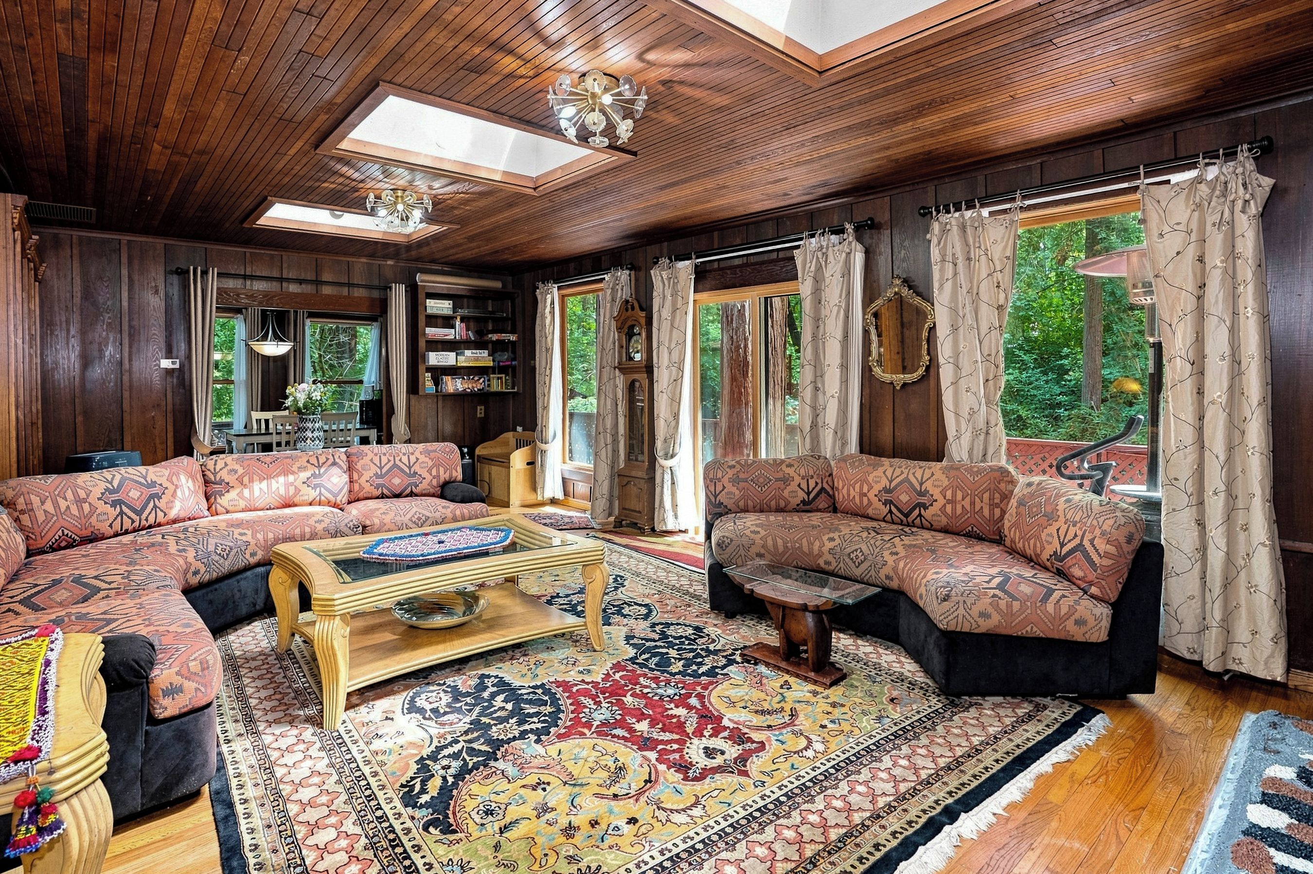 A spacious living room with wooden paneling, a large window with curtains, and a traditional sofa set.