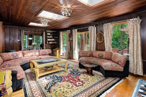 A spacious living room with wooden paneling, a large window with curtains, and a traditional sofa set.