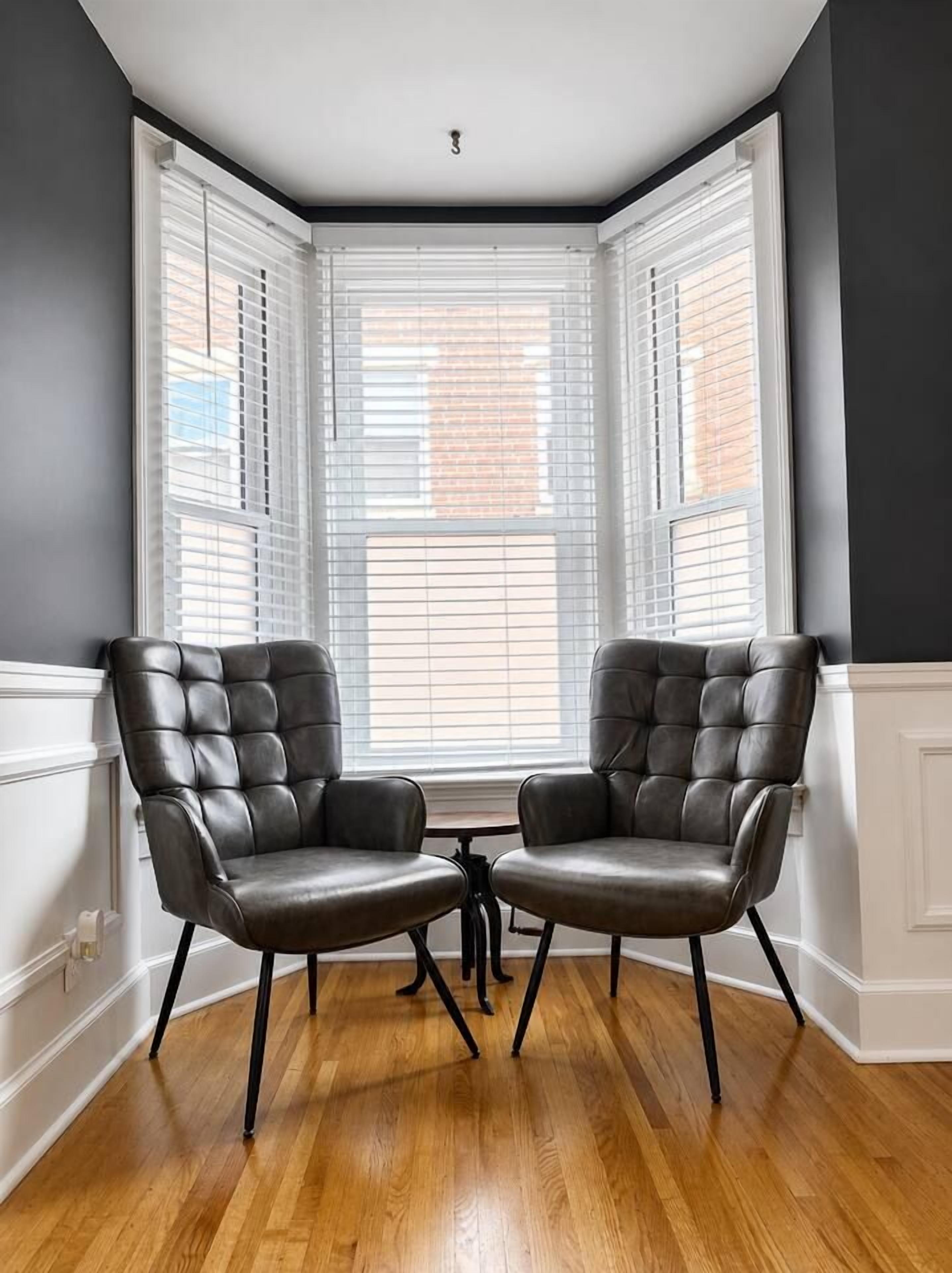 Habitación de esquina con dos sillones de cuero, piso de madera y ventanal con persianas.