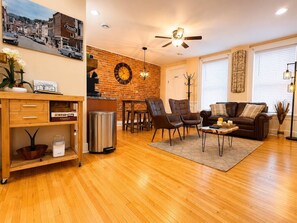 A living room with a wooden floor, a brown leather sofa, a coffee table, and a dining area with a brick wall and a clock. - Suite Victory #5 On Main St w/Reserved Parking (Galena)