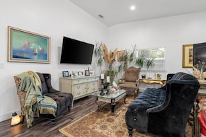 A living room with a dark blue armchair, a beige sofa, a flat-screen TV, a painting, and a decorative plant. - Newly Constructed 1-bedroom guest house near Arden Park (Sacramento)