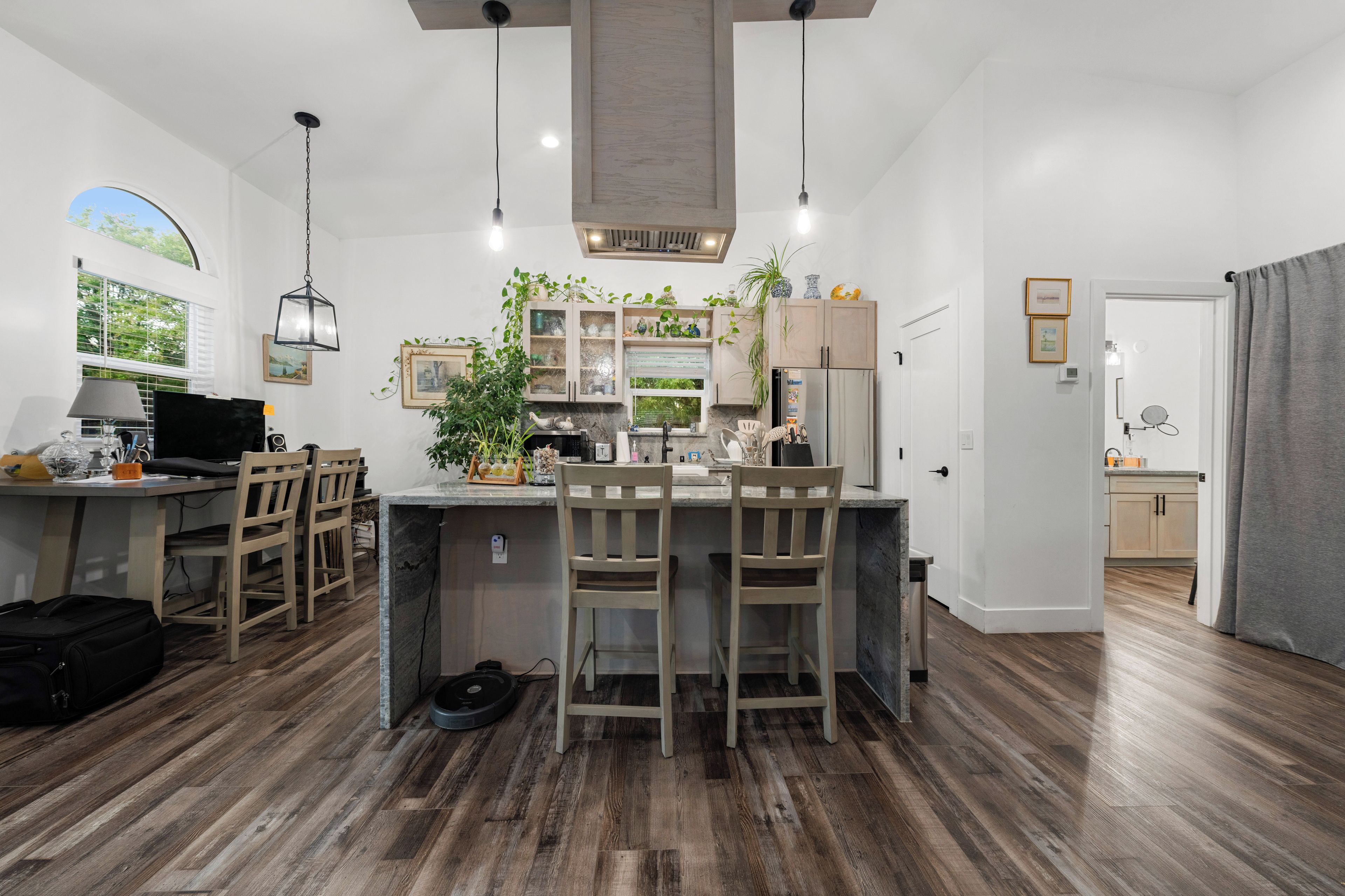 A modern kitchen with wooden flooring, a central island with bar stools, and a view of the outdoors through a window.