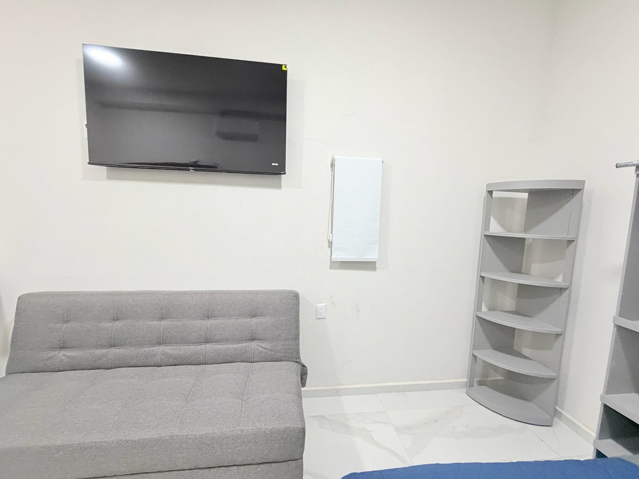 A room with a grey sofa, a TV mounted on the wall, a white wall-mounted shelf, and a corner shelving unit.