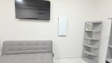 A room with a grey sofa, a TV mounted on the wall, a white wall-mounted shelf, and a corner shelving unit.