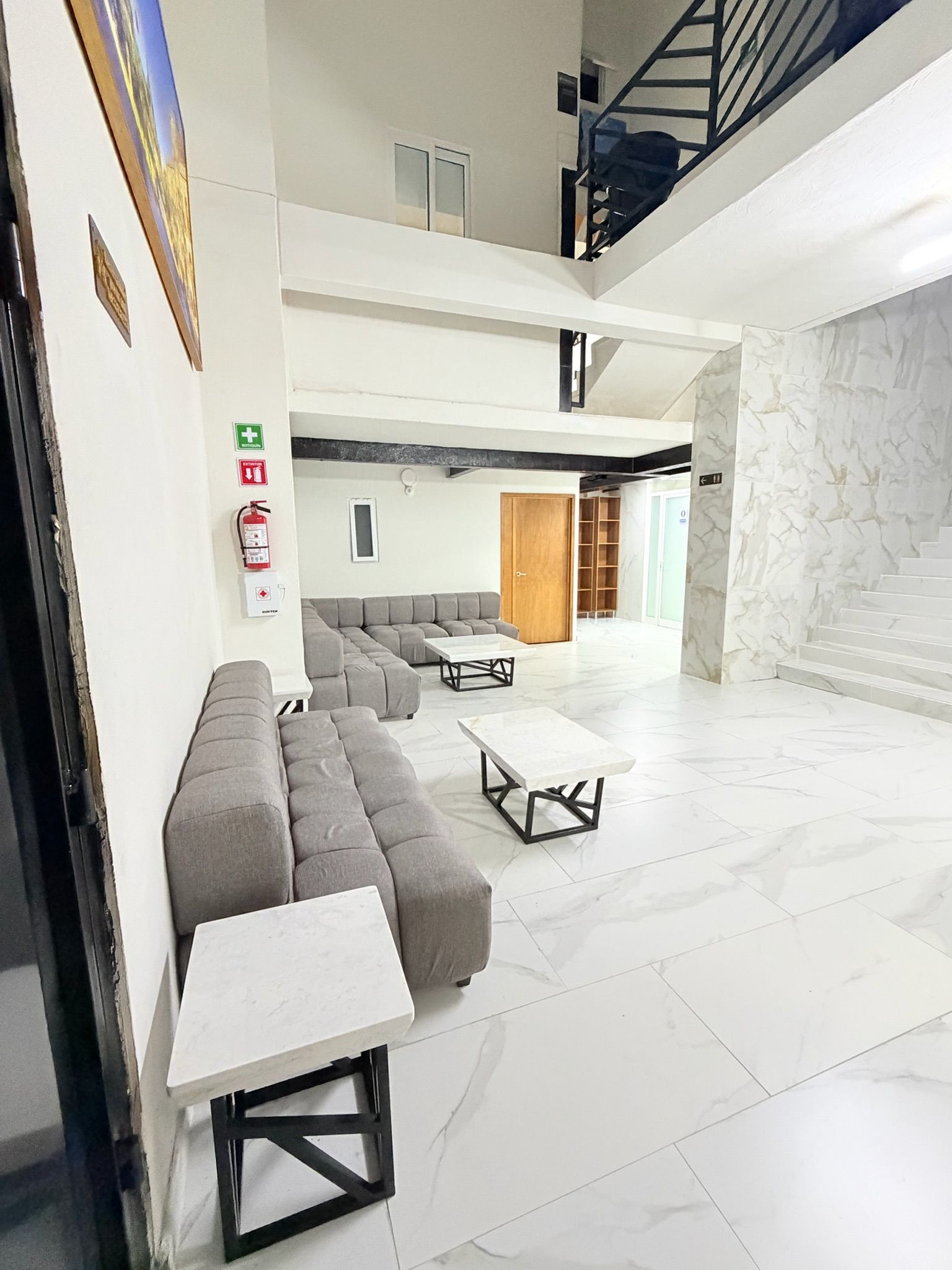 A spacious lobby with a grey sectional sofa, a small white table, a staircase, and a marble wall.