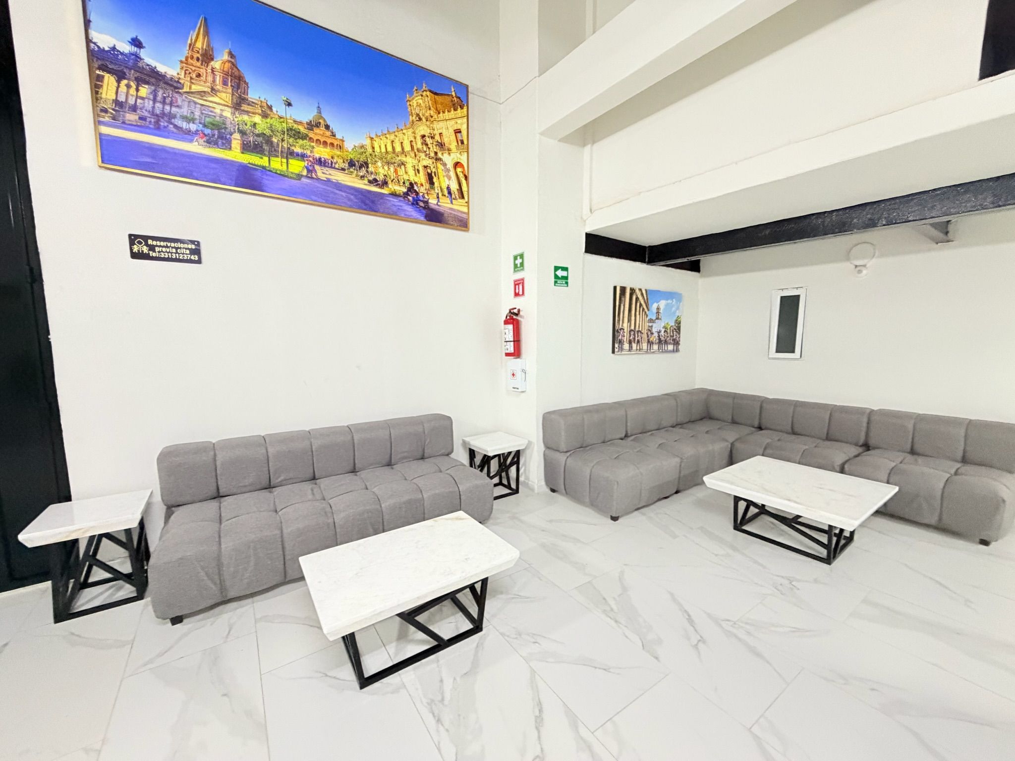 Un área de espera moderna con sofás grises, mesas blancas y un cuadro grande de un edificio histórico.