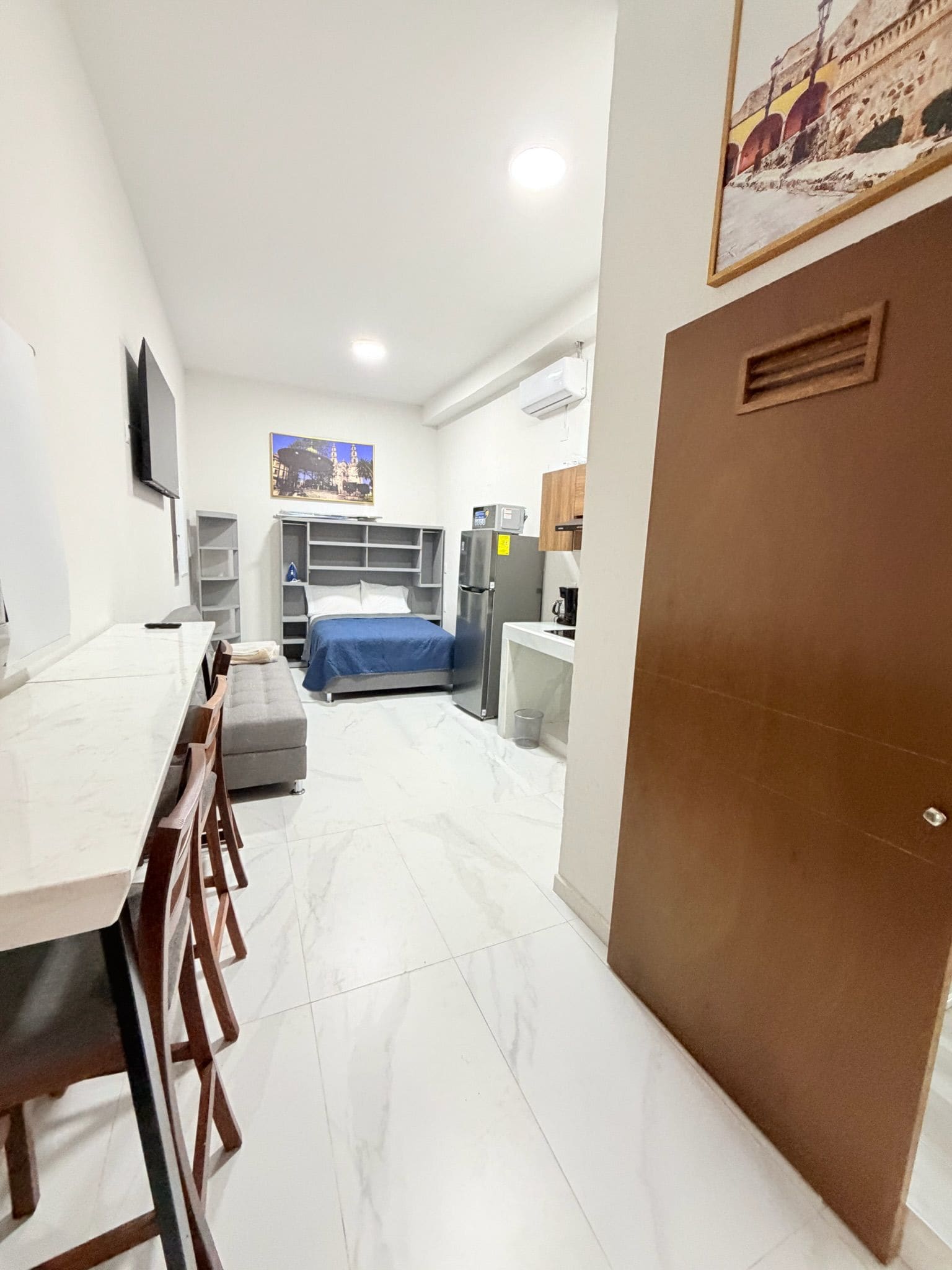 A compact hotel room with a bed, a desk, a chair, a TV, and a kitchen area.